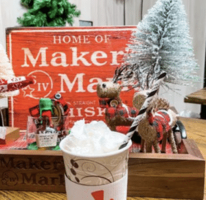 A paper cup of hot chocolate topped with whipped cream sits in front of a wooden tray with decorative reindeer, trees, and a red Maker's Mark sign.